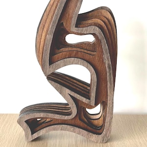 Peut inclure: Sculpture en bois multicouche avec un design abstrait et fluide. L'œuvre présente des tons de bois brun clair et foncé, créant une sensation de profondeur et de dimension. La pièce a une forme organique unique.