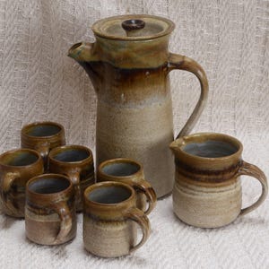 May include: A handcrafted ceramic coffee set. It includes a tall coffee pot with a lid, a creamer jug, and six mugs. The set has a speckled, earthy colour scheme of brown, tan, and blue, with a rustic, handcrafted appearance.