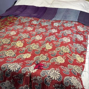 Może przedstawiać: Czerwono-biała kołdra we wzory kwiatowe z motywem paisley. Kołdra jest narzucona na łóżko z białymi poduszkami i fioletowo-niebieskim kocem. Kołdra ma wygląd vintage i jest ręcznie robiona.