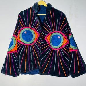May include: A navy blue velvet jacket with embroidered eye designs in orange, pink, blue, and purple. The jacket has a high collar and long sleeves, hanging on a yellow hanger against a white background.
