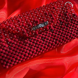 Pode incluir: Uma clutch retangular vermelha com contas com uma etiqueta de metal prateada que diz "ELOURA". A clutch é feita de pequenas contas redondas, aveludadas e contas maiores facetadas. O fundo é um tecido vermelho e sedoso.