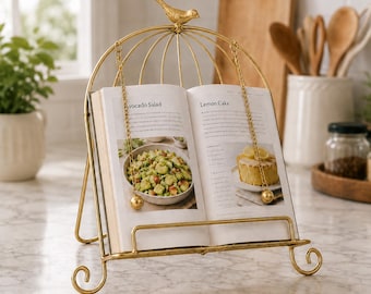 May include: A gold-colored metal cookbook stand featuring a bird-shaped finial at the top and two hanging chains with spherical weights to hold pages open. The stand has a curved wire frame with scroll feet and holds an open cookbook.