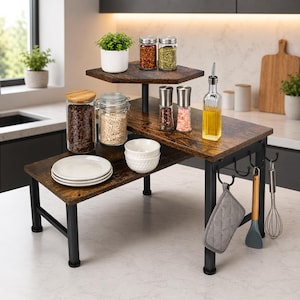 May include: A three-tiered kitchen shelf with a dark wood-look finish and black metal legs. The shelf holds various kitchen items, including spices, plates, bowls, and a bottle of oil. A potholder, spatula, and whisk hang from hooks on the side.