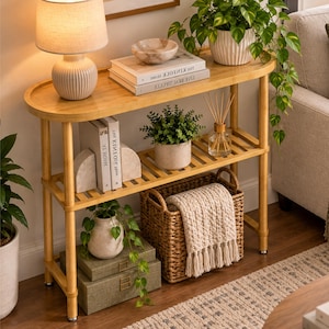 May include: A light wood console table with two tiers, featuring a ribbed table lamp, books titled The Kinfolk Home, a small bowl, potted plants, a reed diffuser, two stacked green storage boxes, and a woven basket with a cream throw blanket.