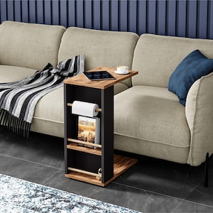 May include: A modern side table with a black frame and a wooden top. It features a built-in magazine rack, a paper towel holder, and a small tabletop. A tablet and a cup of coffee are on the tabletop. The table is next to a beige sofa.
