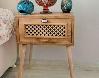 Puede incluir: Una mesita de noche de madera con un cajón y un diseño de celosía en la parte delantera. La mesita tiene tres patas anguladas. Los elementos decorativos en la parte superior incluyen un globo azul, un cuenco decorativo y un jarrón.