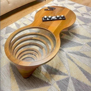 May include: A unique wooden coffee table with a glass-lined, spiral-shaped center. The table has a light brown finish and a distinctive hourglass shape. A small tray with decorative items sits on the table's surface. The table is set on a patterned rug.