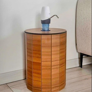 Op de afbeelding: Een cilindervormige houten bijzettafel met een waterdispenser. De tafel is gemaakt van verticale houten panelen in bruintinten. De waterdispenser is wit en grijs met een zilveren tuit. Een blauwe waterfles is zichtbaar.
