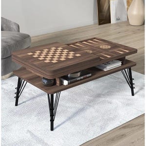 Peut inclure: Table basse en bois brun foncé avec un échiquier et un jeu de backgammon intégrés. La table a des pieds en épingle à cheveux en métal noir et une étagère inférieure pour le rangement. La table est sur un tapis blanc.