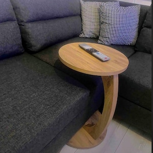 May include: A small, oval-shaped wooden side table with a curved leg design. The table is light brown and holds a remote control. It sits next to a dark gray sectional sofa with two houndstooth pattern pillows.