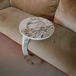 May include: A circular side table with a white marble top and a curved marble base. The marble features grey veining. The table is placed beside a beige sofa.