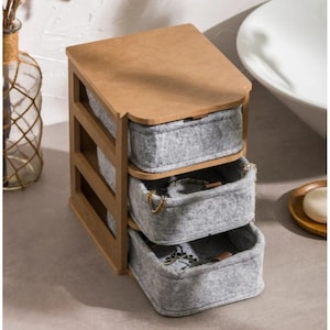 Puede incluir: Un organizador de joyas de madera con tres cajones forrados de fieltro gris. La parte superior y el marco son de madera marrón claro. Los cajones están parcialmente abiertos, revelando joyas en su interior. El organizador está diseñado para colocarse sobre una encimera.