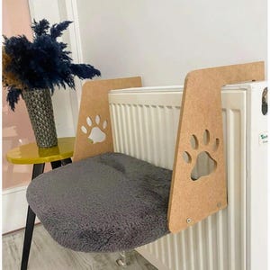 Könnte beinhalten: Ein Katzen-Heizkörperbett mit einem flauschigen grauen Kissen. Der Holzrahmen hat Pfotenabdruck-Ausschnitte. Das Bett ist so konzipiert, dass es an einem Heizkörper aufgehängt werden kann und einen warmen, gemütlichen Platz zum Entspannen bietet.