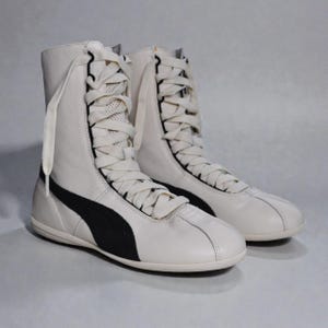 May include: Pair of white leather high-top sneakers with black side stripes and long white laces. The sneakers have a padded collar and a rubber sole. The Puma logo is visible on the side.