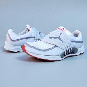 May include: Pair of white and gray athletic shoes with a red sole. The shoes feature a mesh upper, a gray strap with a Puma logo, and gray accents. The Puma logo is also visible on the tongue.
