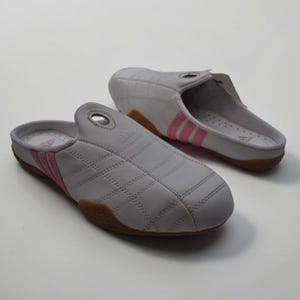 May include: Light grey Adidas slip-on shoes with pink stripes and brown soles. The shoes feature a quilted pattern and the Adidas logo. These comfortable slip-ons are suitable for casual wear. The soles are brown.
