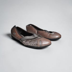 May include: A pair of brown leather ballet flats. The shoes have a rounded toe and a decorative panel with small perforations. A gray elastic band crosses the top of the foot. The soles are black.