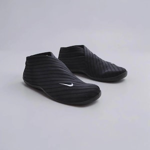 May include: A pair of black slip-on shoes featuring a textured, wavy striped pattern across the upper material. Each shoe displays a small, white embroidered Nike swoosh logo on the outer side near the toe area, with a low-cut ankle design.