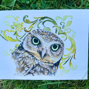 May include: A watercolour painting of an owl with green eyes, brown and white feathers, and a yellow and green floral design in the background.