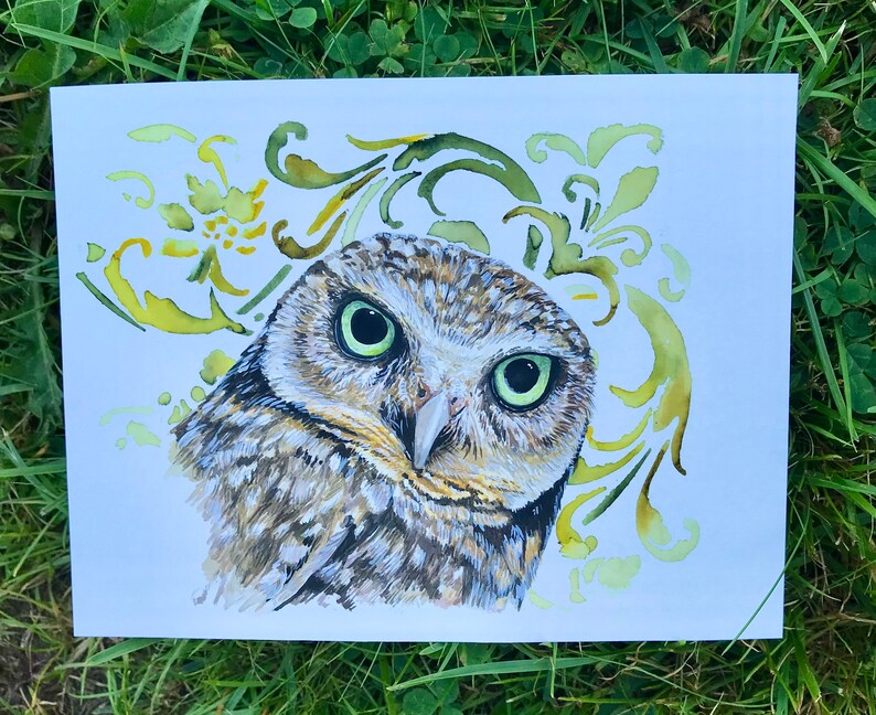 May include: A watercolour painting of an owl with green eyes, brown and white feathers, and a yellow and green floral design in the background.