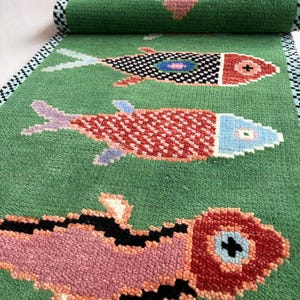 Könnte beinhalten: Grüner Teppich mit pixeligem Fischdesign in Rot, Schwarz, Rosa und Blau. Die Fische sind auf einem grünen Hintergrund angeordnet, mit einem schwarz-weißen Rand. Gerollte Teppiche sind im Hintergrund zu sehen.