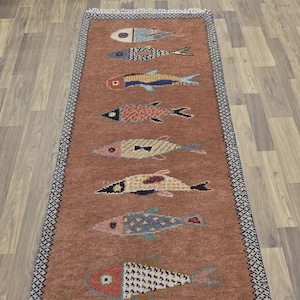 Op de afbeelding: Een rechthoekig, terracotta-kleurig tapijt met een herhaald patroon van gestileerde vissen in verschillende kleuren. De visontwerpen omvatten blauw, geel, rood en groen, met gedetailleerde patronen. Het tapijt heeft een donkerblauwe rand met een herhalend geometrisch ontwerp.