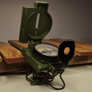 May include: An open, olive-green military-style compass with a black sighting arm. The compass features a rotating dial with directional markings and a ruler along the side. The compass is resting on a wooden surface.