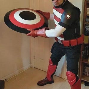 Puede incluir: Un disfraz negro y rojo con un escudo con un dise&ntilde;o blanco, rojo y negro. El atuendo incluye un casco, guantes y botas negros. El disfraz tiene un emblema de estrella en el hombro.