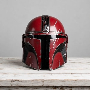 Op de afbeelding: Een rood en zwart helm met een verweerde uitstraling, die doet denken aan een Mandalorian helm. De helm heeft een afgeronde bovenkant met een zwarte streep en een hoekig frontpaneel. De helm staat op een wit houten oppervlak.