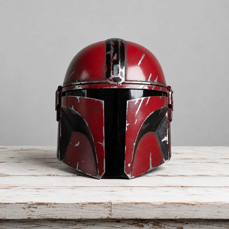 Op de afbeelding: Een rood en zwart helm met een verweerde uitstraling, die doet denken aan een Mandalorian helm. De helm heeft een afgeronde bovenkant met een zwarte streep en een hoekig frontpaneel. De helm staat op een wit houten oppervlak.
