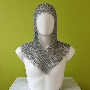 Peut inclure: Une coiffe en cotte de mailles, un couvre-chef médiéval, exposée sur un buste de mannequin blanc. La coiffe est faite de petits anneaux métalliques imbriqués, créant une couche protectrice flexible. Le fond est vert citron.