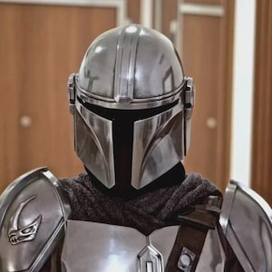 Op de afbeelding: Een zilveren Mandalorian helm en harnasset. De helm heeft een T-vormig vizier en verticale ribbels. De bepantsering omvat schouderstukken met een gestileerd embleem en een borstplaat. Een donkere stof bedekt de nek.