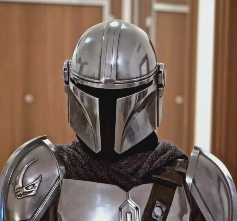 Op de afbeelding: Een zilveren Mandalorian helm en harnasset. De helm heeft een T-vormig vizier en verticale ribbels. De bepantsering omvat schouderstukken met een gestileerd embleem en een borstplaat. Een donkere stof bedekt de nek.