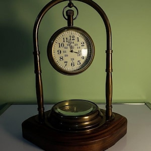 May include: A vintage desk clock with a round clock face and a brass-coloured frame. The clock has a dark wooden base and a magnifying glass. The clock face displays the numbers 1 to 12 and 13 to 24.