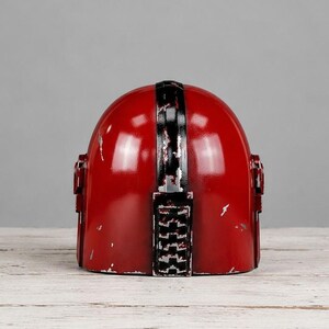 Op de afbeelding: Een rode helm met een zwarte streep in het midden en zwarte accenten. De helm heeft een verweerde afwerking, met zichtbare slijtage. Het ontwerp doet denken aan een ruimtethema.
