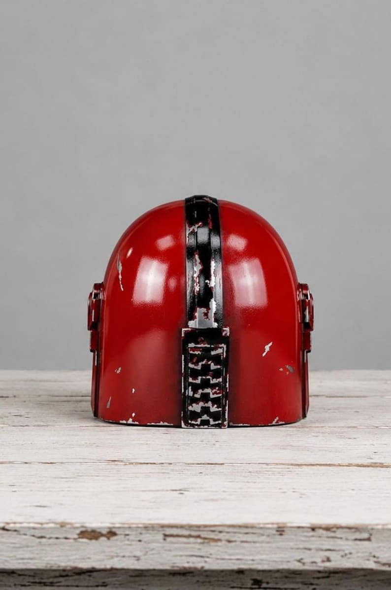 Op de afbeelding: Een rode helm met een zwarte streep in het midden en zwarte accenten. De helm heeft een verweerde afwerking, met zichtbare slijtage. Het ontwerp doet denken aan een ruimtethema.