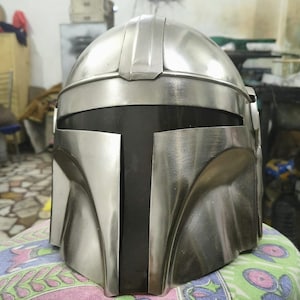 Op de afbeelding: Een zilverkleurige Mandalorian helm met een afgeronde bovenkant en een T-vormig vizier. De helm heeft een metallic afwerking en is ontworpen met hoekige kenmerken. Het viziergedeelte is zwart, wat contrasteert met de zilveren buitenkant.