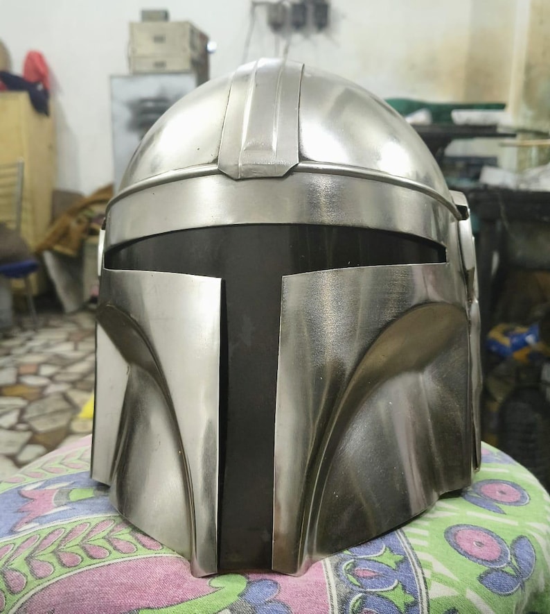 Op de afbeelding: Een zilverkleurige Mandalorian helm met een afgeronde bovenkant en een T-vormig vizier. De helm heeft een metallic afwerking en is ontworpen met hoekige kenmerken. Het viziergedeelte is zwart, wat contrasteert met de zilveren buitenkant.