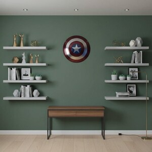 K&ouml;nnte beinhalten: Ein Raum mit einer dunkelgr&uuml;nen Wand und wei&szlig;en schwebenden Regalen. Die Regale zeigen B&uuml;cher, kleine Pflanzen und geometrische Skulpturen. Ein Captain America-Schild h&auml;ngt an der Wand, und ein Holzschreibtisch mit schwarzen Beinen befindet sich darunter.