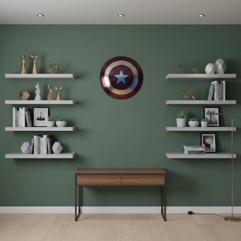 K&ouml;nnte beinhalten: Ein Raum mit einer dunkelgr&uuml;nen Wand und wei&szlig;en schwebenden Regalen. Die Regale zeigen B&uuml;cher, kleine Pflanzen und geometrische Skulpturen. Ein Captain America-Schild h&auml;ngt an der Wand, und ein Holzschreibtisch mit schwarzen Beinen befindet sich darunter.