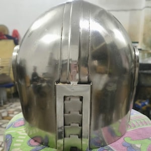 Op de afbeelding: Een gepolijste, zilverkleurige helm met een afgeronde bovenkant en verticale ribbels. De achterkant van de helm heeft een rechthoekig, gesegmenteerd ontwerp. De helm lijkt van metaal te zijn en reflecteert de omgeving.