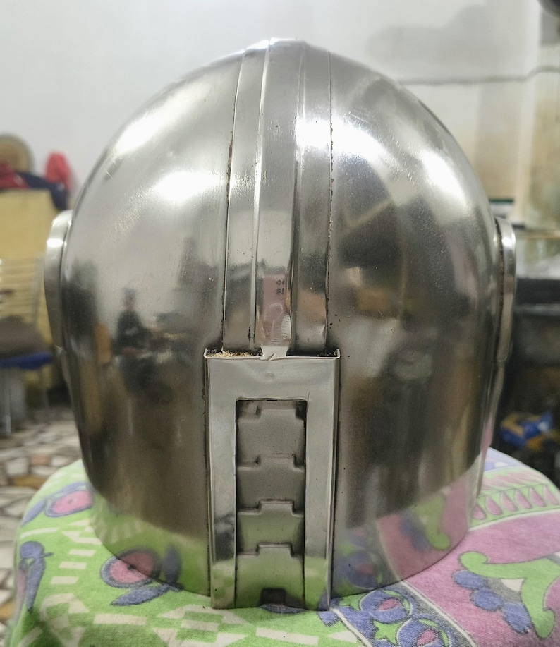 Op de afbeelding: Een gepolijste, zilverkleurige helm met een afgeronde bovenkant en verticale ribbels. De achterkant van de helm heeft een rechthoekig, gesegmenteerd ontwerp. De helm lijkt van metaal te zijn en reflecteert de omgeving.