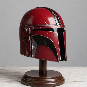 Op de afbeelding: Een rode en zwarte Mandalorian helm replica, gepresenteerd op een houten standaard. De helm heeft een distressed afwerking, met zwarte accenten en een vizier. De standaard heeft een ronde basis en een donkerbruine afwerking.
