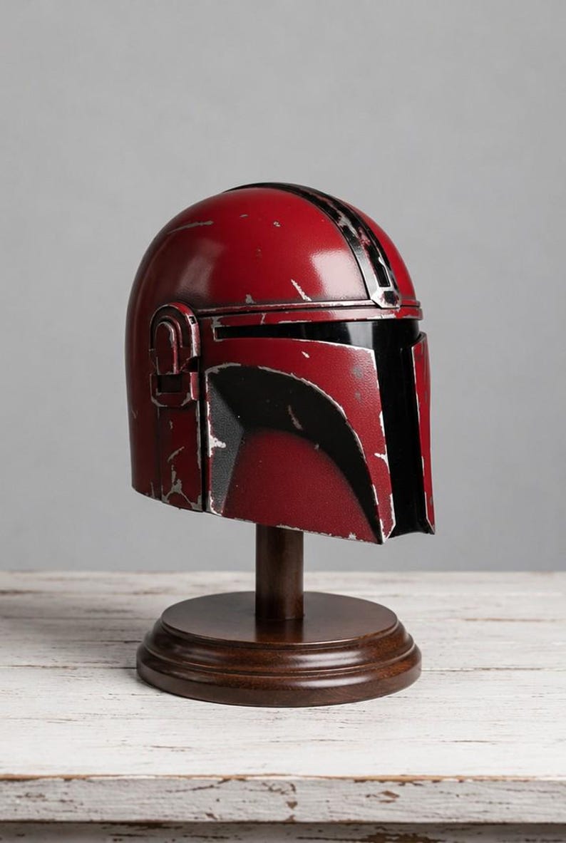 Op de afbeelding: Een rode en zwarte Mandalorian helm replica, gepresenteerd op een houten standaard. De helm heeft een distressed afwerking, met zwarte accenten en een vizier. De standaard heeft een ronde basis en een donkerbruine afwerking.