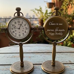 May include: Two antique brass-coloured pocket watch clocks. One clock displays the time, the other has the inscription "Cheers to new journeys, life awaits. Happy Birthday". Each clock is supported by a decorative stand.