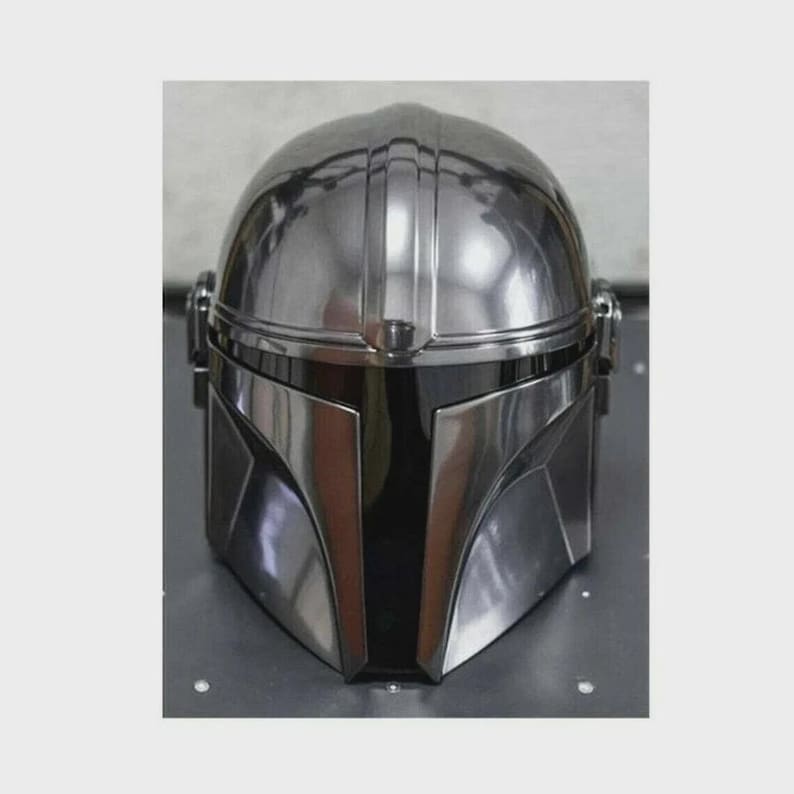 Op de afbeelding: Een glanzende, zilverkleurige Mandalorian helm met een opvallend T-vormig vizier. De helm heeft een afgeronde bovenkant met verticale groeven en een gladde, metalen afwerking. Het ontwerp doet denken aan sciencefiction.
