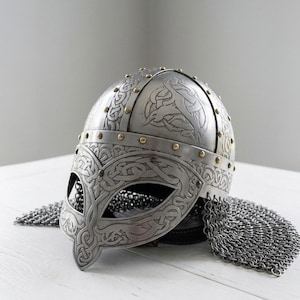 Peut inclure: Un casque viking argenté avec des motifs gravés complexes et des rivets en laiton. Le casque est doté d'une protection faciale et d'une protection de cou en cotte de mailles. Le design rappelle l'artisanat nordique historique.