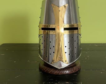 May include: A silver and gold medieval crusader helmet with a cross design. The helmet has a polished metal finish with a gold cross and accents. It is mounted on a dark wooden base, showcasing historical armor.