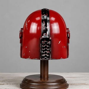 Op de afbeelding: Een rode helm met zwart-witte details, gepresenteerd op een houten standaard. De helm vertoont tekenen van slijtage, met een verweerde afwerking. De standaard is donkerbruin en de achtergrond is neutraal grijs.