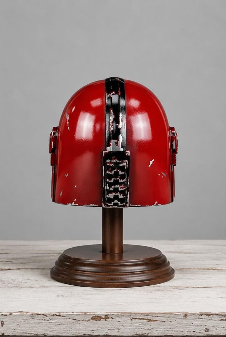 Op de afbeelding: Een rode helm met zwart-witte details, gepresenteerd op een houten standaard. De helm vertoont tekenen van slijtage, met een verweerde afwerking. De standaard is donkerbruin en de achtergrond is neutraal grijs.